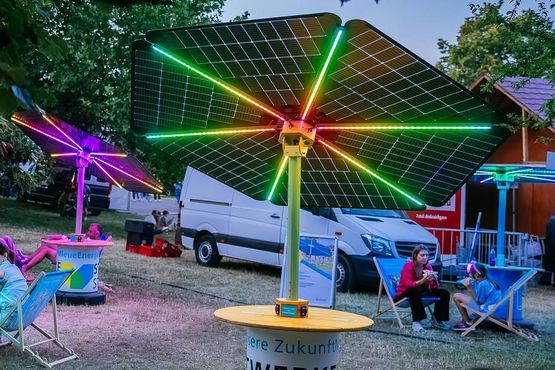Mehrere Solar-Sonnenschirme der Stadtwerke Stuttgart stehen in der Dämmerung auf einer Wiese und sind an der Unterseite mit LED beleuchtet.