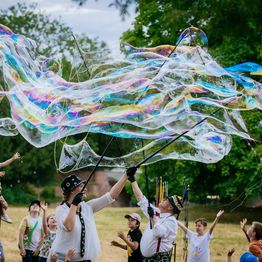 Frohnella Klücklich vom Clowntheater macht auf einer Wiese vor zahlreichen Kindern riesige Seifenblasen