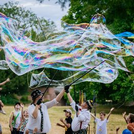 Frohnella Klücklich vom Clowntheater macht auf einer Wiese vor zahlreichen Kindern riesige Seifenblasen